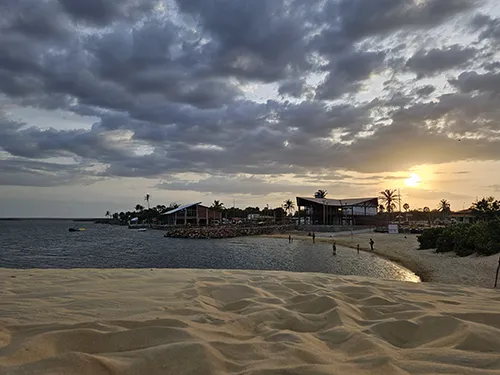 Pôr do sol na Lagoa do Portinho, Parnaíba, PI, visto das dunas de areia, com restaurantes modernos à beira da água e céu com nuvens dramáticas.