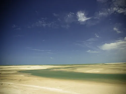 Paisagem ampla da Praia de Macapá em Luís Correia, PI, na maré baixa, mostrando vasta extensão de areia molhada com canais de água rasa (espelhos d'água) sob um céu azul com poucas nuvens.