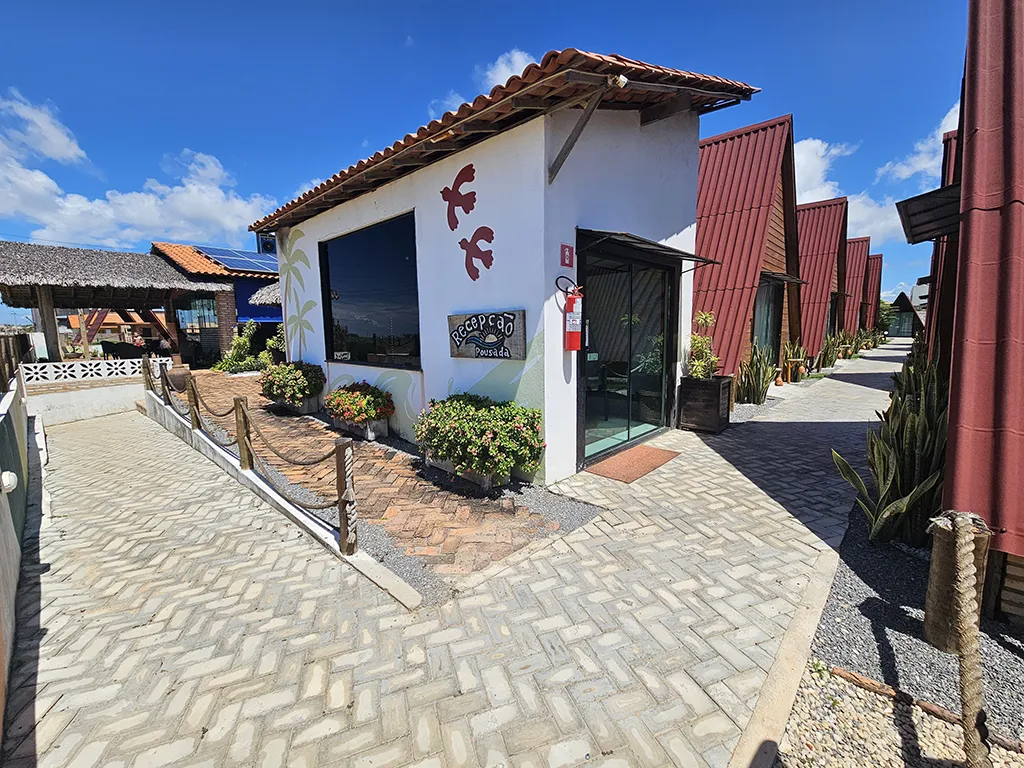 Recepção dos Chalés Al Mare: Sua Chegada ao Paraíso em Luís Correia Vista externa da área da recepção dos Chalés Al Mare, mostrando a entrada do prédio branco com detalhes em vermelho e madeira, e o caminho para os chalés.